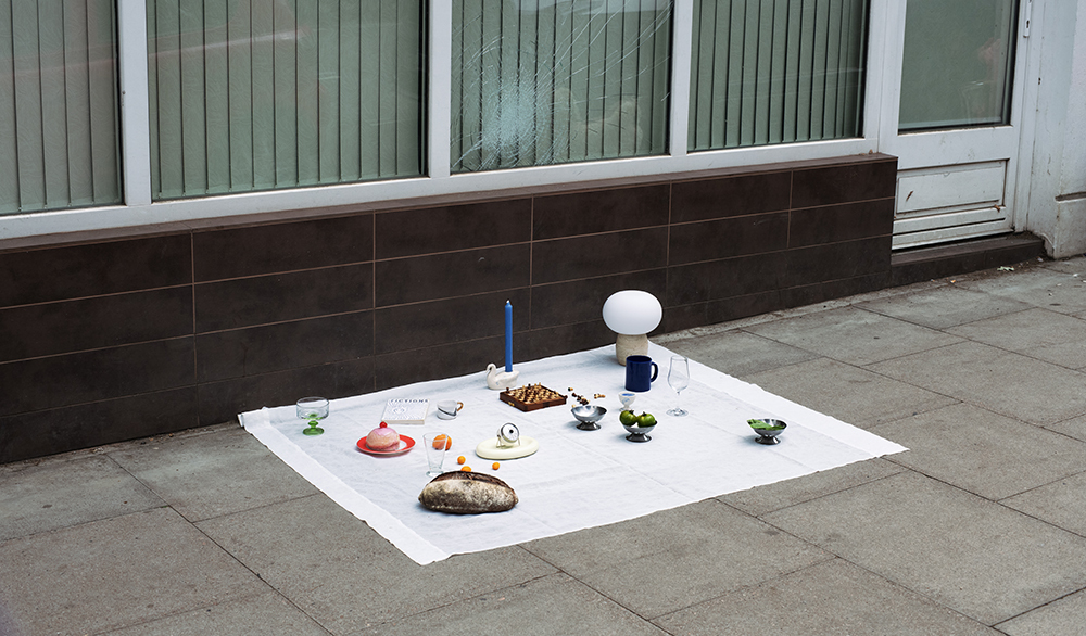 A picnic on a white blanket on the pavement outside of a solicitor's office. The window of the office is broken.